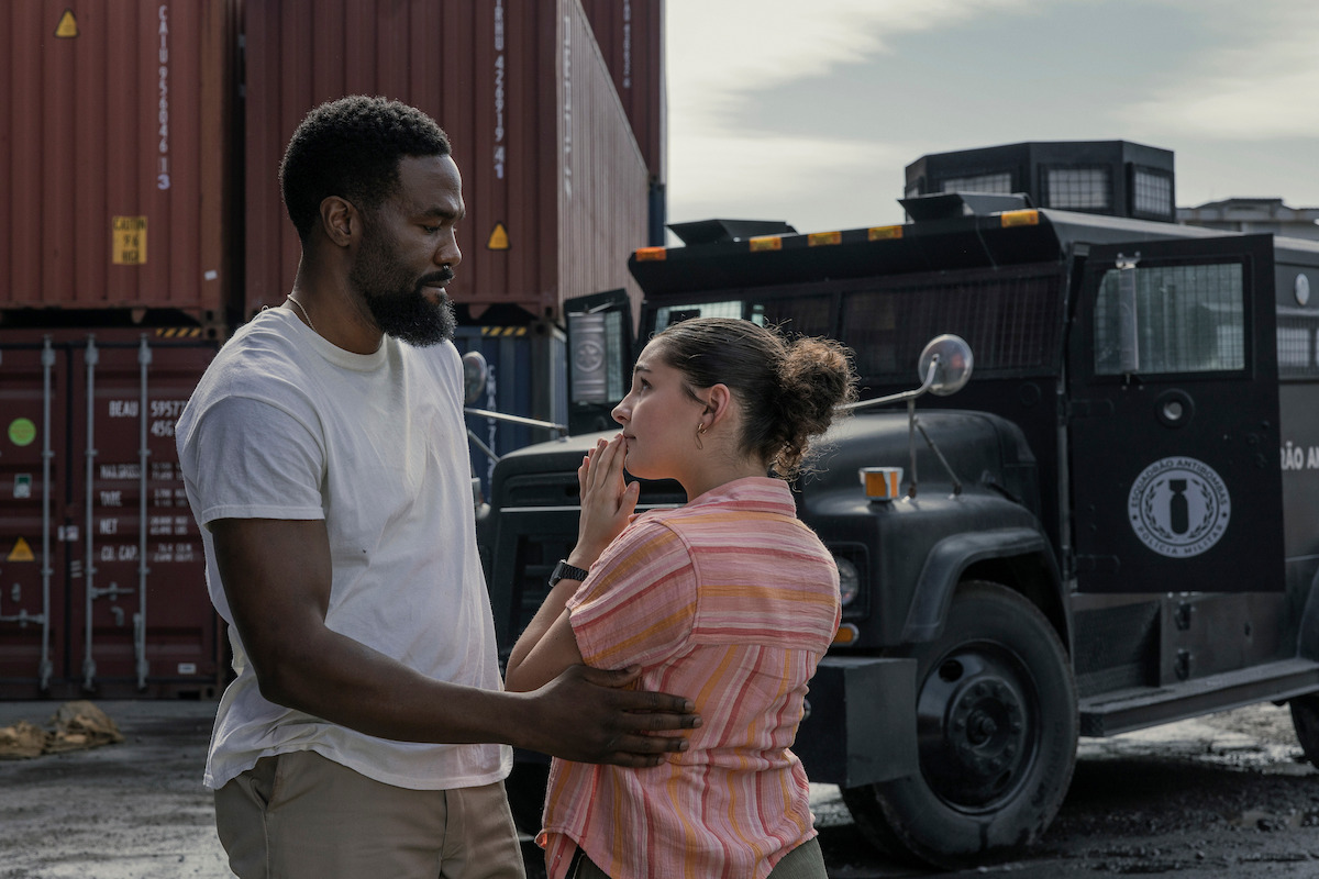 A man comforts a concerned woman outside near shipping containers and an armored truck in an industrial setting.