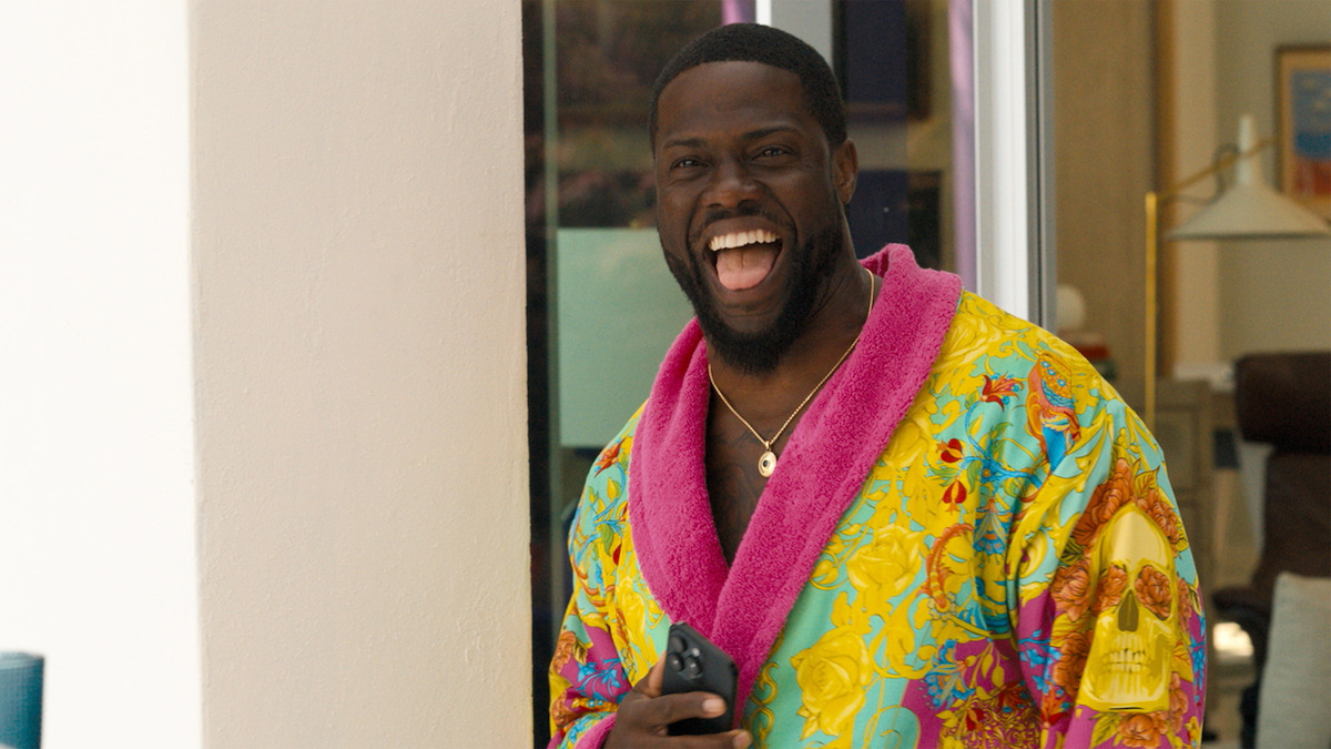 Man standing indoors near a doorway, laughing joyfully, wearing a colorful bathrobe with a bright pink collar and holding a smartphone, with modern home furnishings in the background.