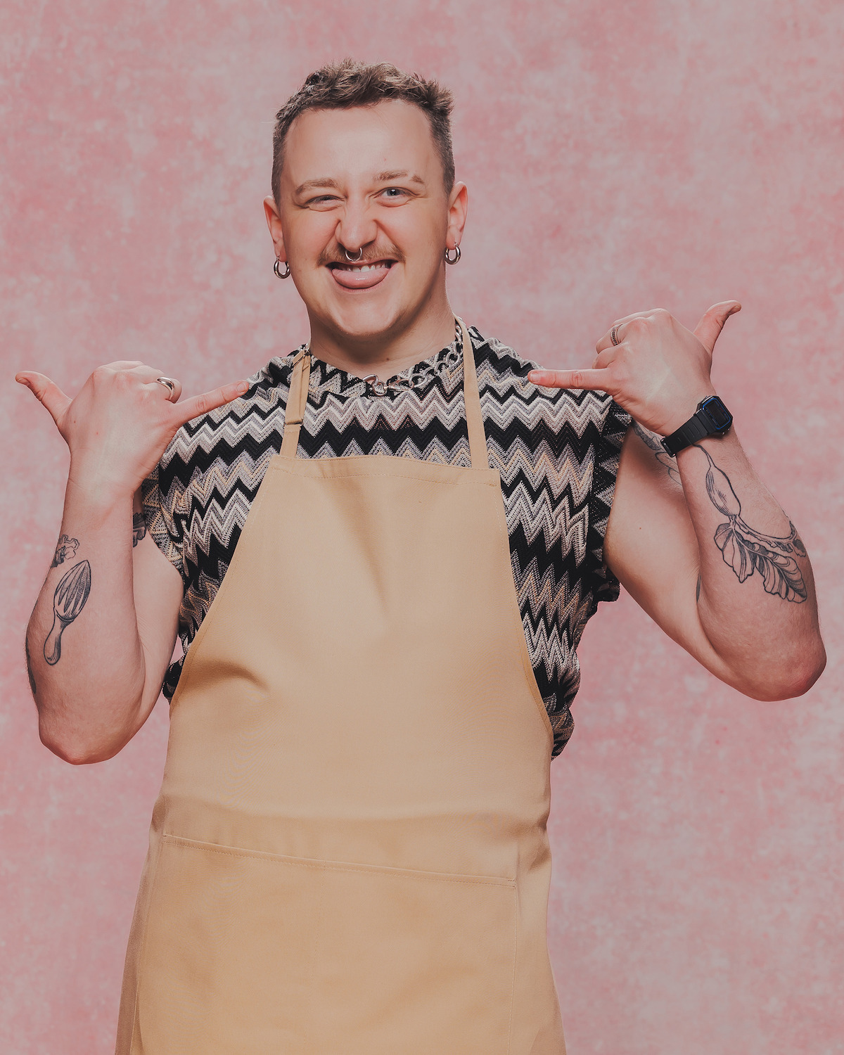 Person with short hair, tattoos, and piercings wearing a beige apron and geometric patterned shirt, smiling and pointing at themselves in front of a textured pink background in a portrait studio setting.