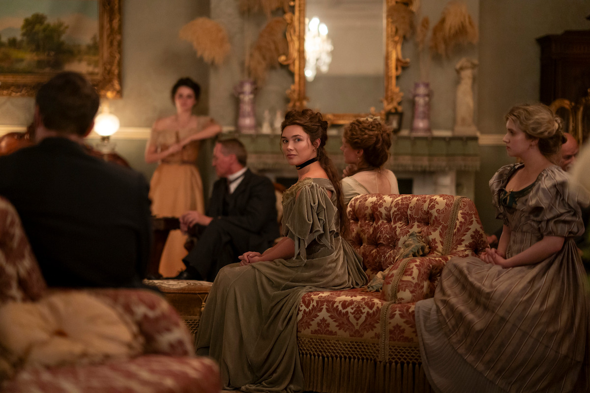 A group of people in period clothing sit in an ornate, vintage living room with elegant furniture, antique decorations, and a large mirror above the fireplace, engaged in conversation or listening.