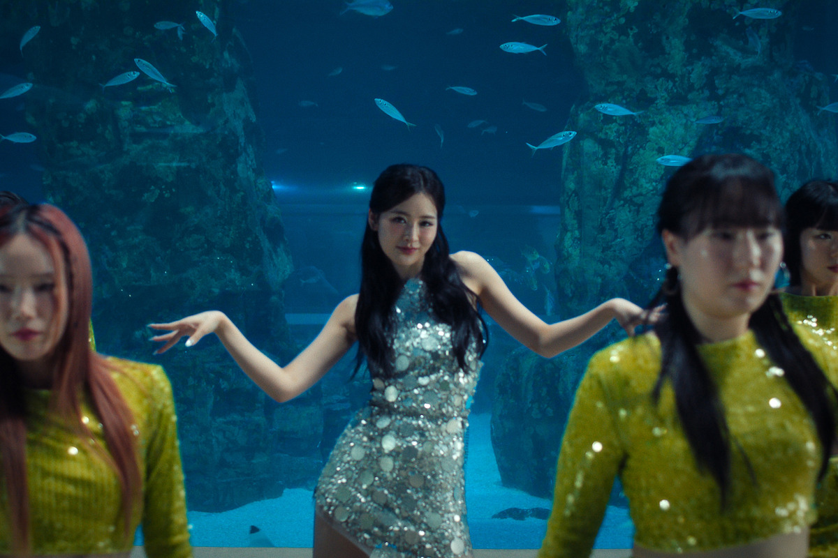 A group of women dancing in a large aquarium setting, with a woman in a sparkly silver dress in the center and dancers in yellow outfits, surrounded by rocks and swimming fish in the background.