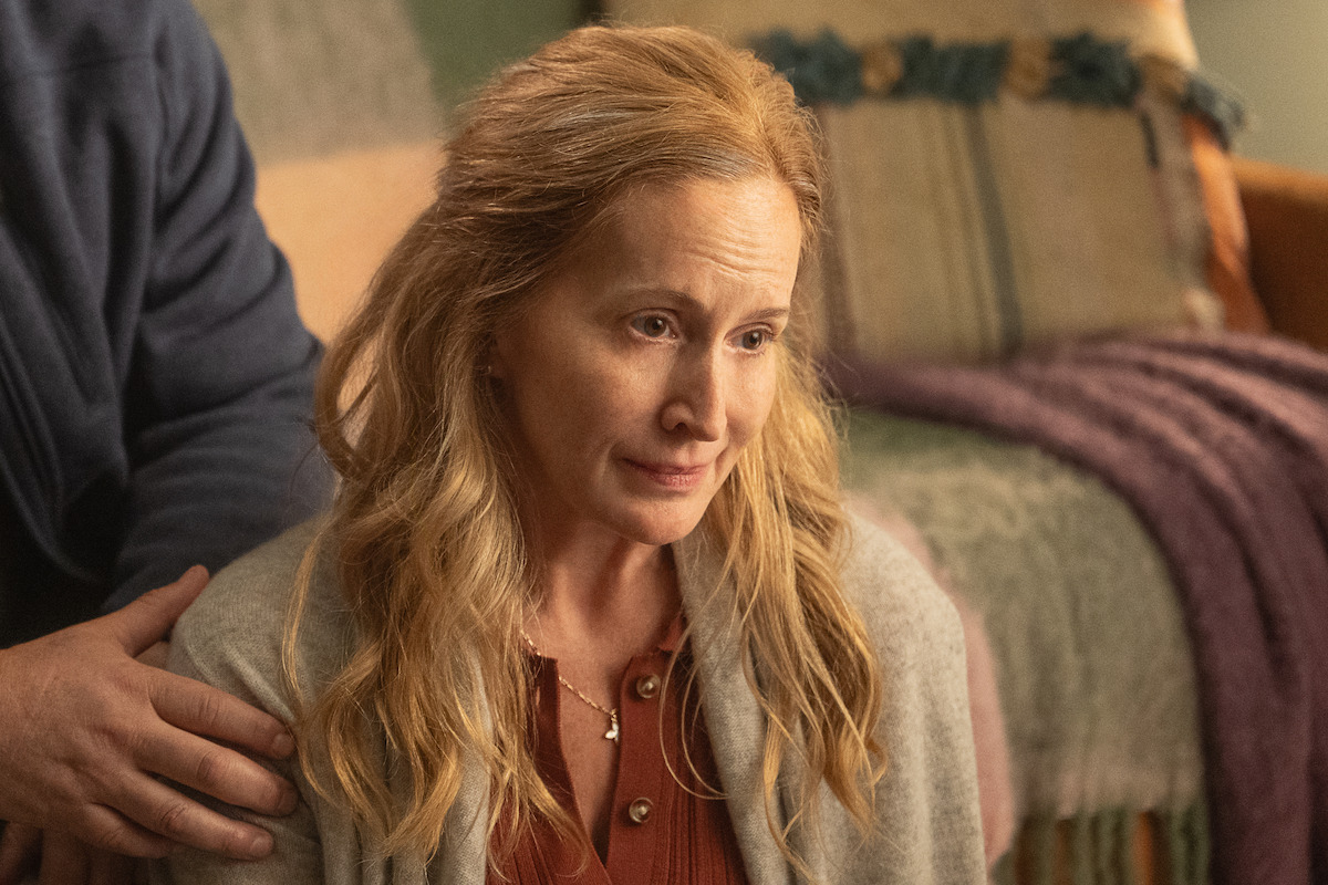 A woman with long blonde hair sits indoors, looking thoughtful or emotional, with a man’s comforting hand on her shoulder. The setting includes a cozy couch and soft blankets in warm lighting.