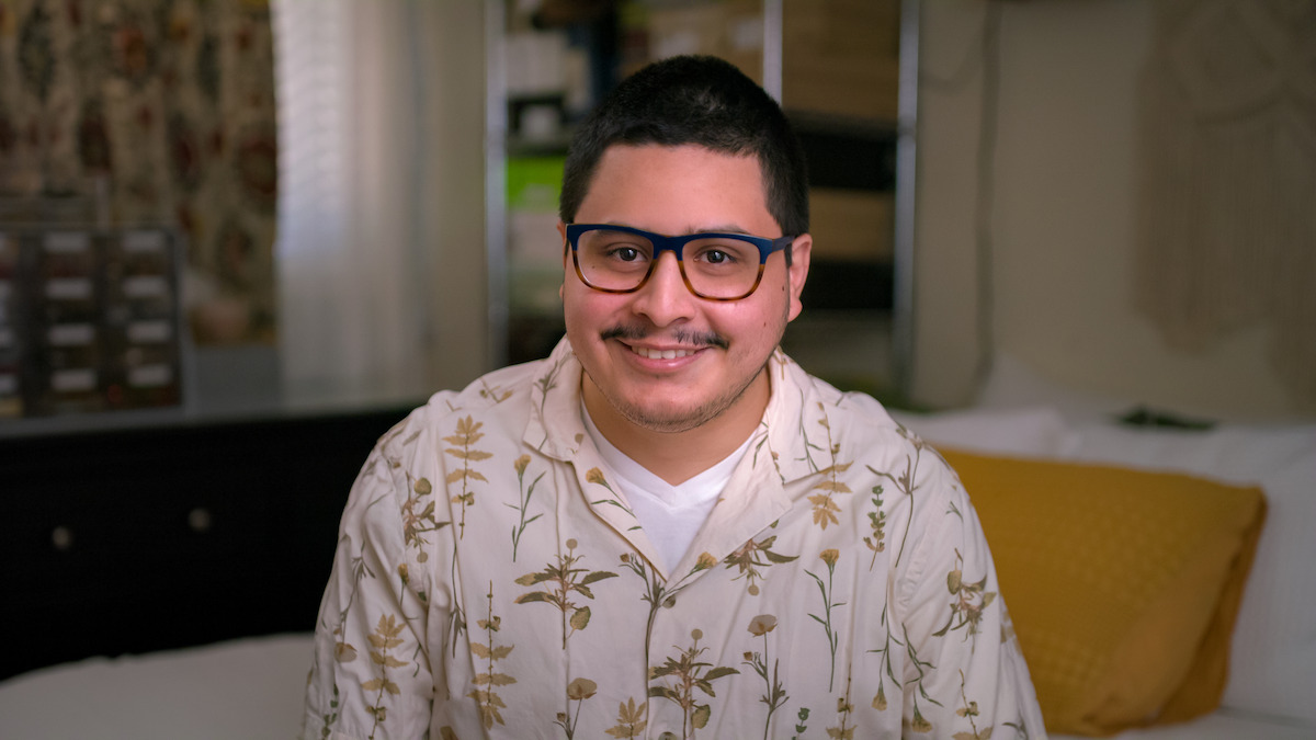 Man with glasses and short hair smiling, wearing a patterned shirt, sitting on a bed in a cozy, softly-lit bedroom with a yellow pillow and furniture in the background.