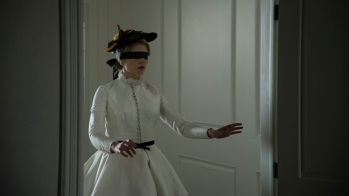 A woman in a white period costume and feathered hat, blindfolded, cautiously steps forward with arms outstretched inside a dimly lit room with white paneled doors.