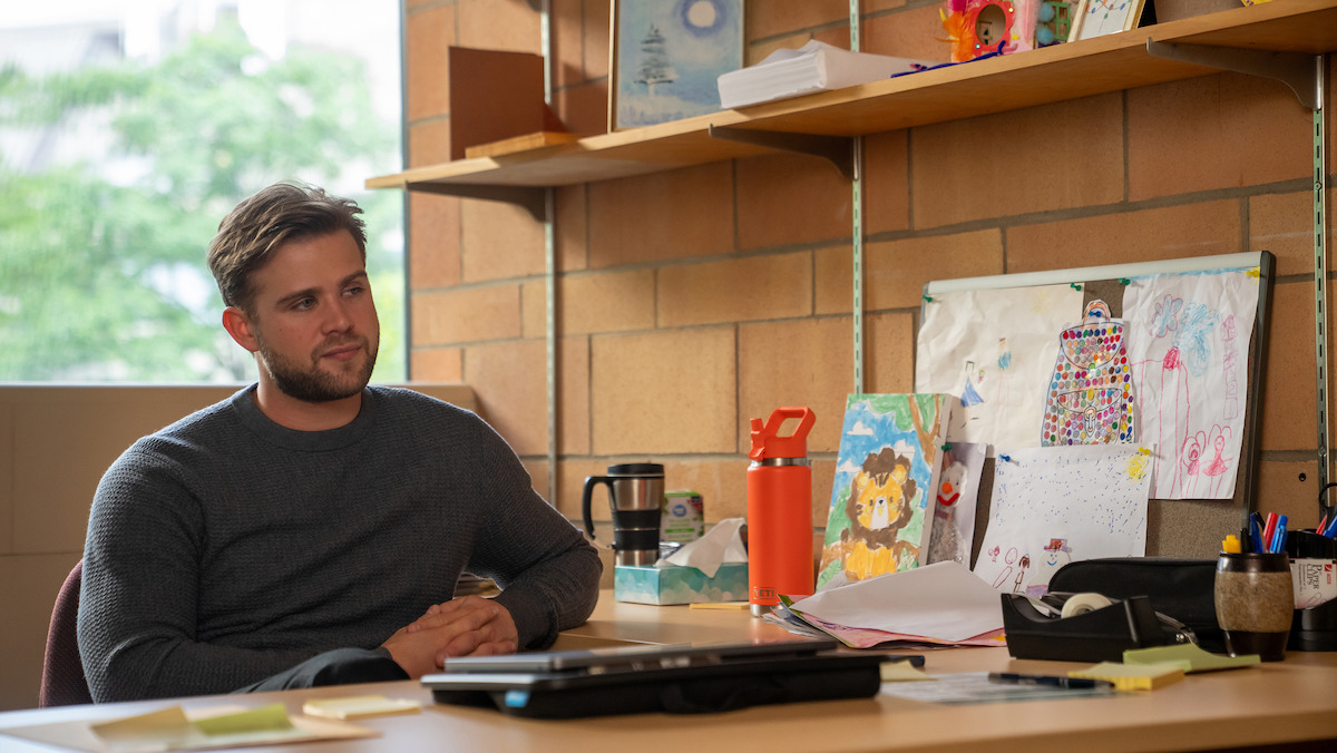 Leo Woodall as Vladimir sits at a desk in an office with brick walls, children's artwork, papers, and office supplies, looking thoughtful. There are shelves with personal items and a window letting in daylight.Leo Woodall as Vladimir sits at a desk in an office with brick walls, children's artwork, papers, and office supplies, looking thoughtful. There are shelves with personal items and a window letting in daylight.