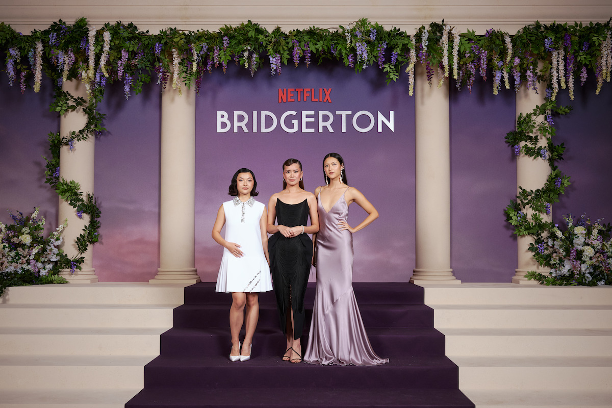 Three women in elegant dresses pose on purple carpeted steps with columns, floral decorations, and a purple backdrop featuring the Netflix Bridgerton logo at a formal event.