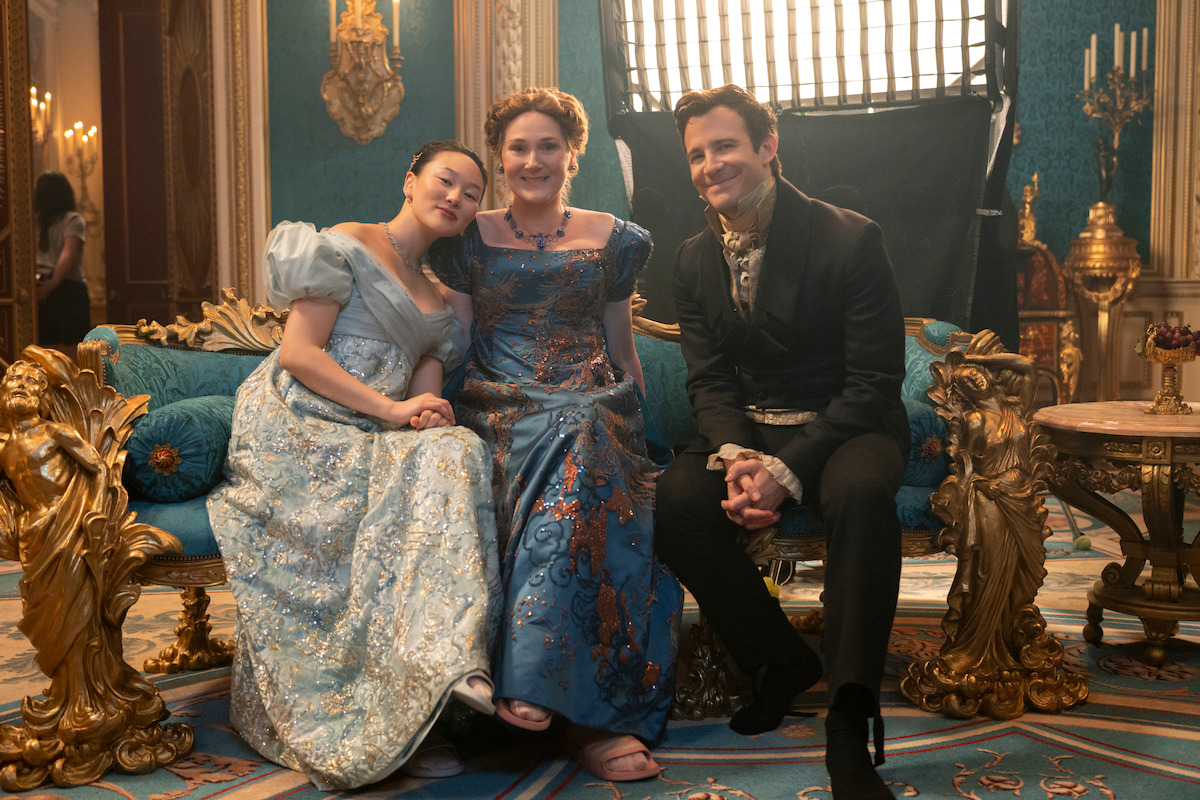 Three people in ornate period costumes sit on a lavish blue and gold settee in an elegant, royally decorated room with blue walls, chandeliers, gold accents, and a film light in the background.