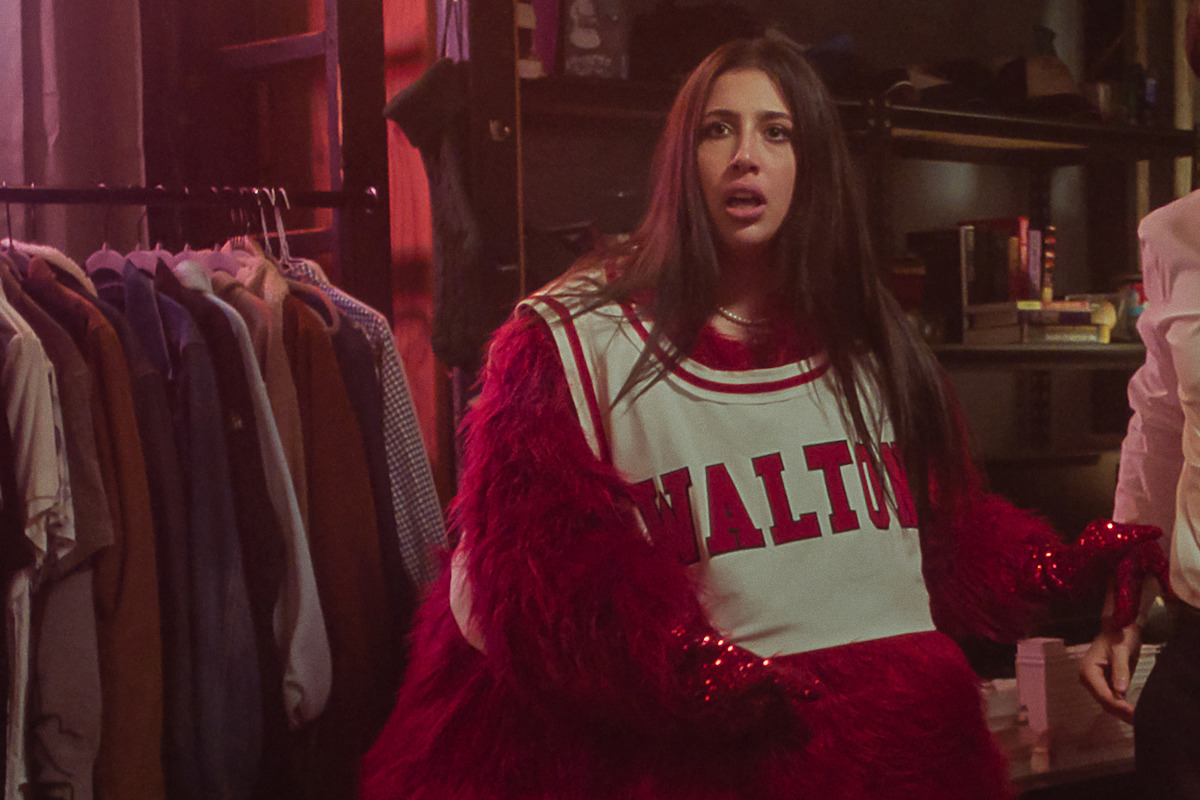 A woman wearing a furry red costume and a jersey stands in a dimly lit room with hanging clothes and shelves in the background, looking surprised or confused.