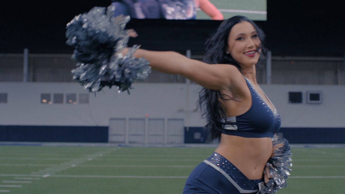 Cheerleader in a blue uniform dances with pom-poms on an indoor football field, smiling and mid-performance, with stadium seating and a scoreboard in the background.Cheerleader in a blue uniform dances with pom-poms on an indoor football field, smiling and mid-performance, with stadium seating and a scoreboard in the background.Cheerleader in a blue uniform dances with pom-poms on an indoor football field, smiling and mid-performance, with stadium seating and a scoreboard in the background.