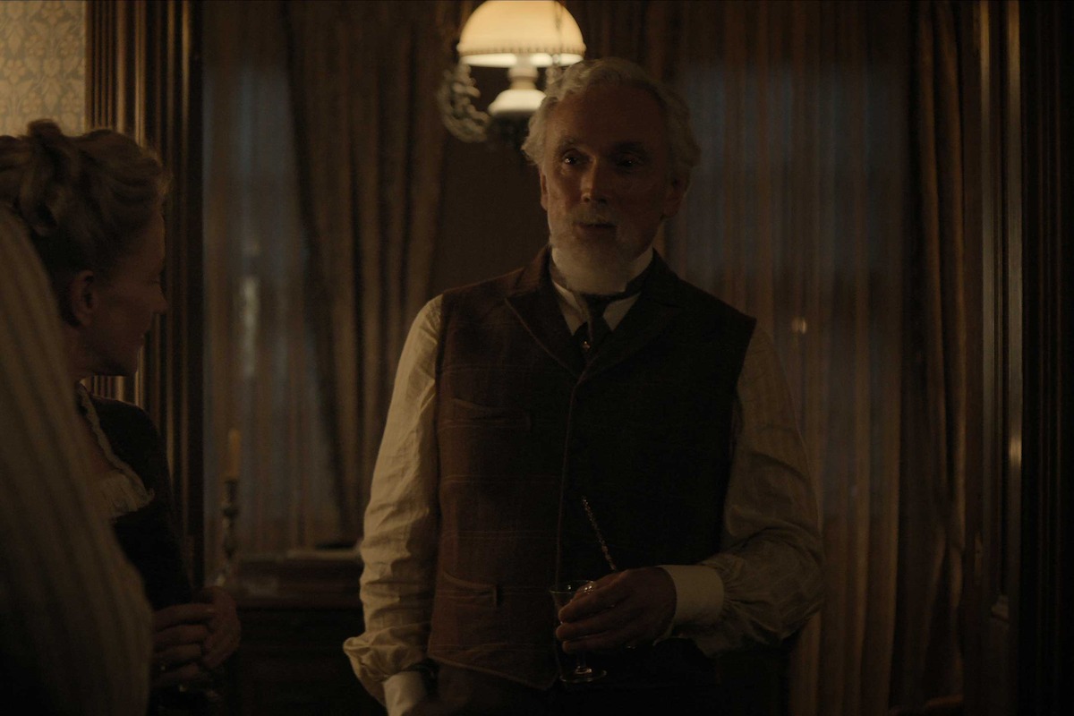 An older man in period clothing stands in a dimly lit vintage room with curtains, holding a drink, facing a woman, creating a somber and contemplative mood.
