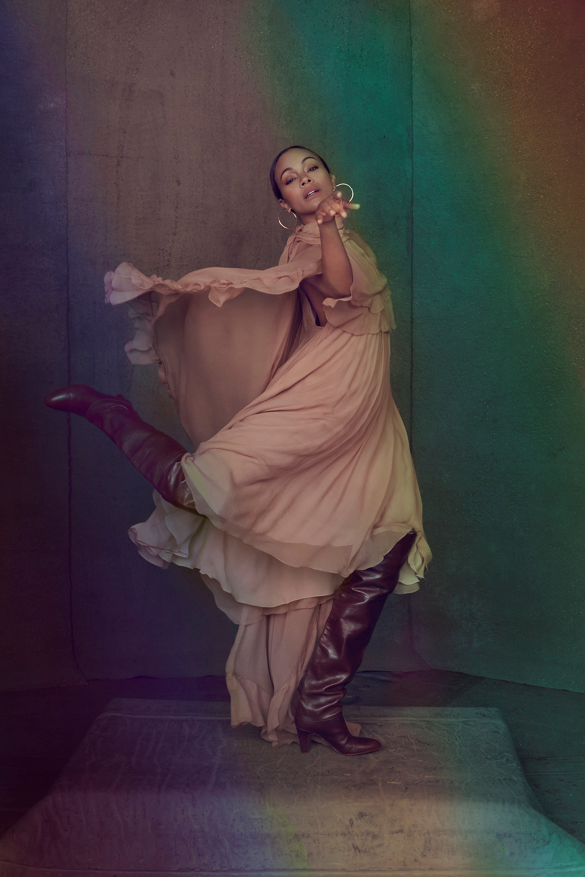 Zoe Saldaña wears a tan dress and brown boots and strikes a pose against a green and gray background.