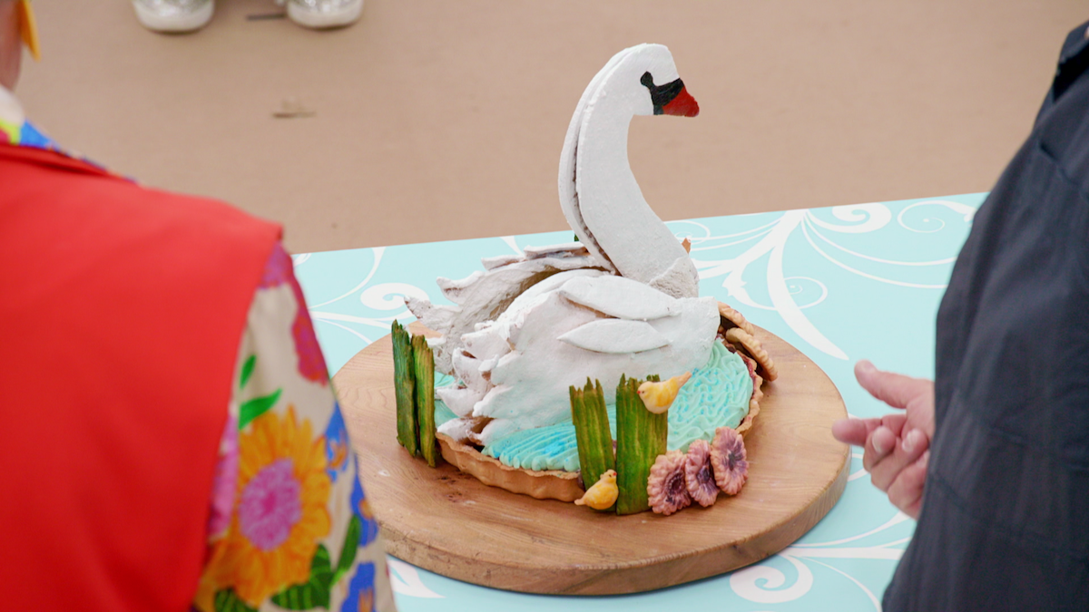 A swan-shaped cake with white frosting, green decorations, and blue icing sits on a wooden platter on a decorated table. Two people stand nearby, one in colorful clothing and another in black. Indoor setting, contest or showcase.