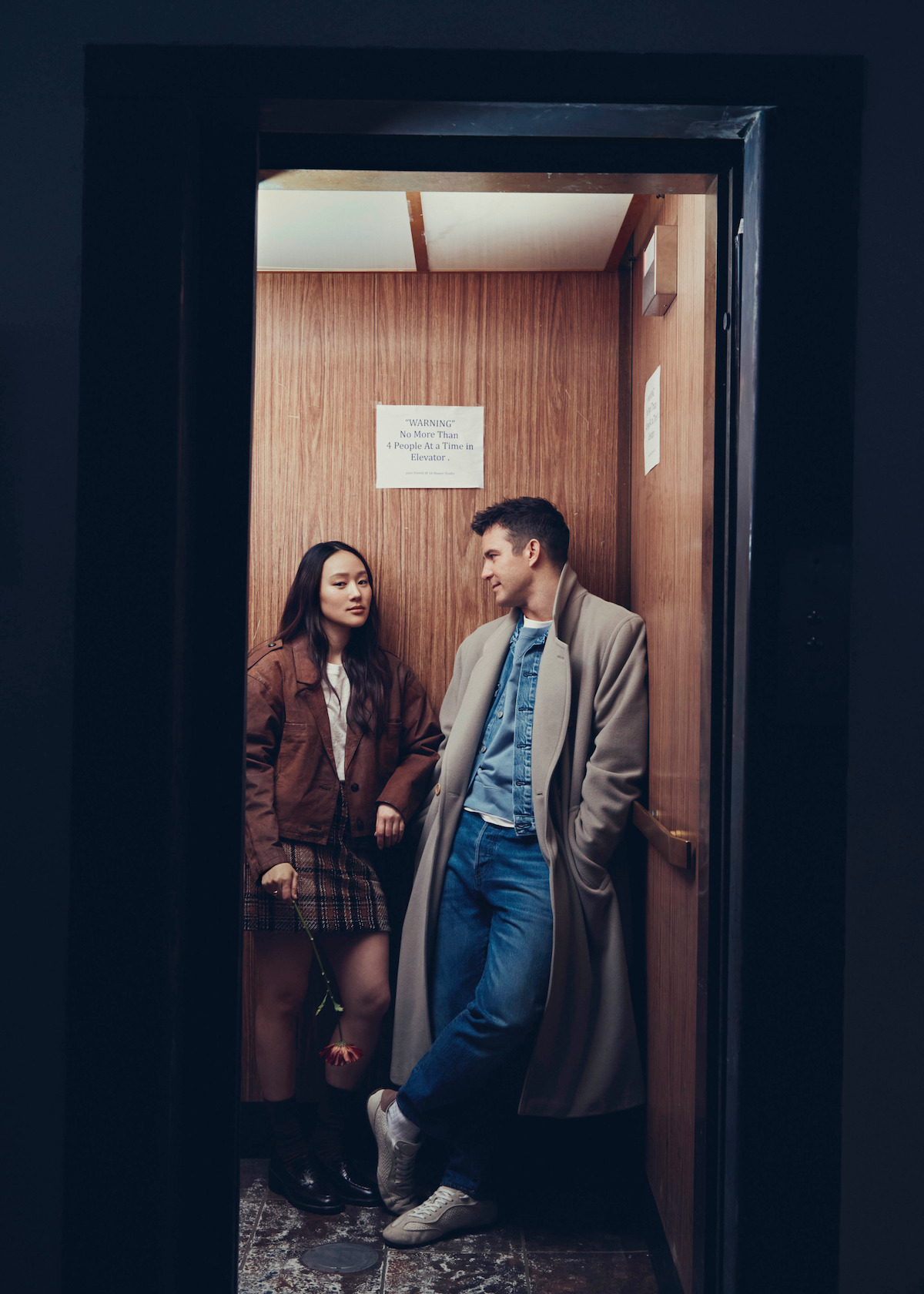 Two people standing in a small, wood-paneled elevator. One holds a rose, both wear coats and casual clothes, and they appear to be talking or waiting together in a dimly lit, intimate setting.