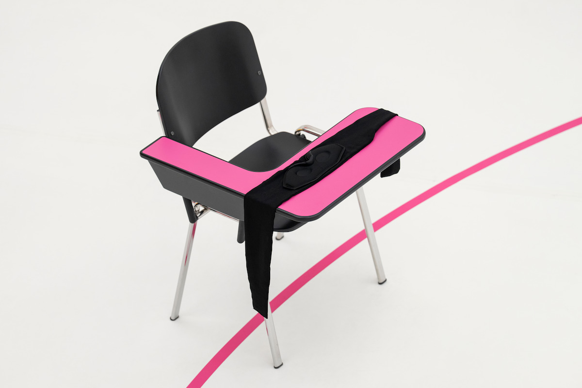 A black chair with an attached desk painted pink, draped with a black cloth, sits on a white floor intersected by a bright pink curved line, creating a modern and minimalist atmosphere.