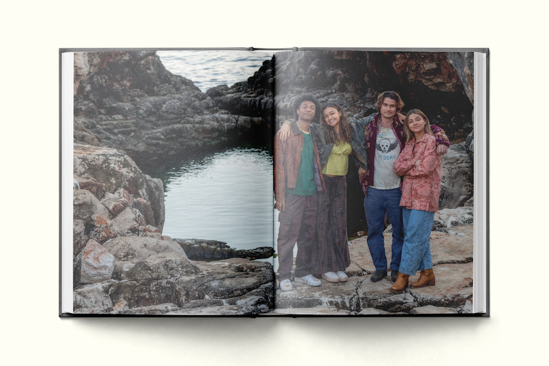 Four young adults stand together smiling on rocky terrain near a small body of water, surrounded by natural rock formations, appearing relaxed and happy outdoors, in a photo printed in an open book.