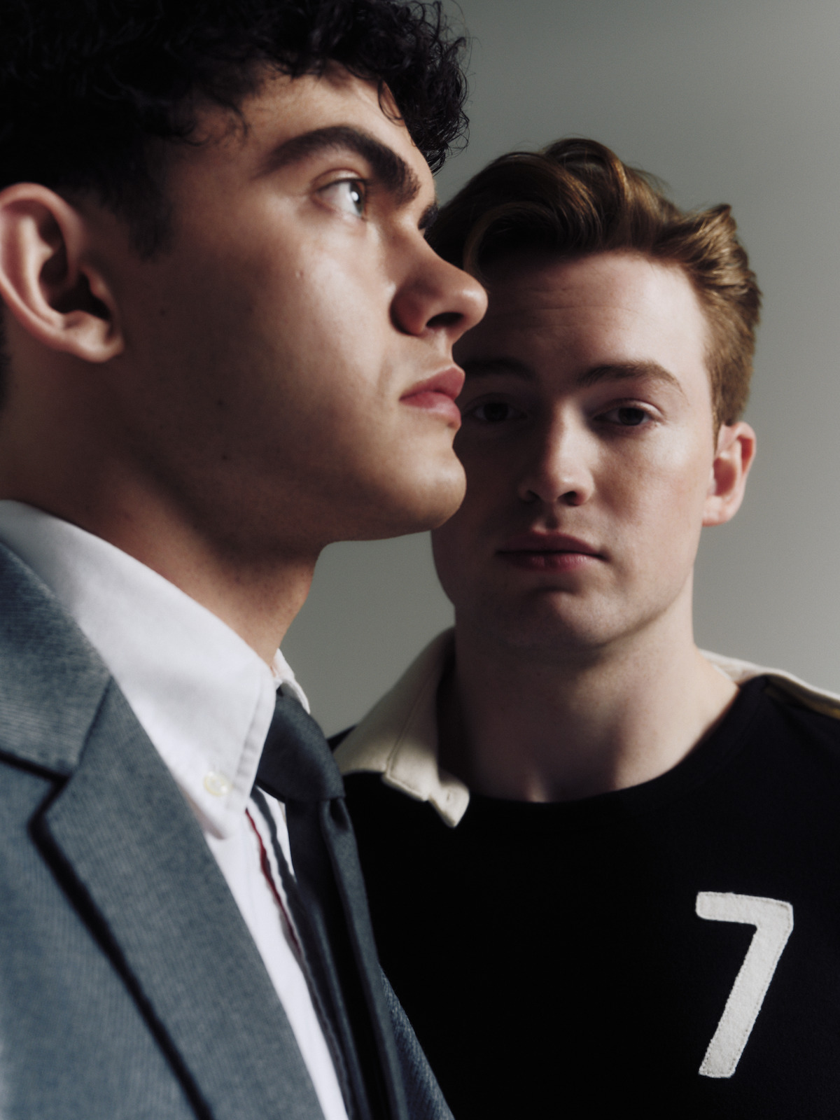 Two young men indoors, one in a gray suit and tie in the foreground looking left, the other in a black shirt with a white number 7 in the background looking at the camera, neutral gray background, soft lighting.