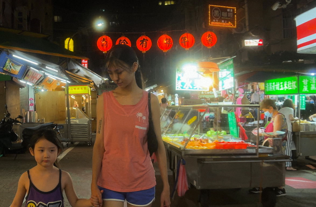 A woman and a child walk through a lively night market with bright lights, food stalls, and red lanterns overhead, creating a warm, bustling atmosphere.