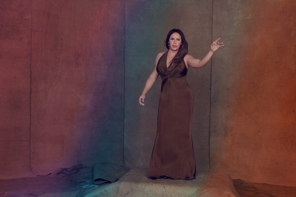 Karla Sofía Gascón wears a long brown dress and raises her arm.