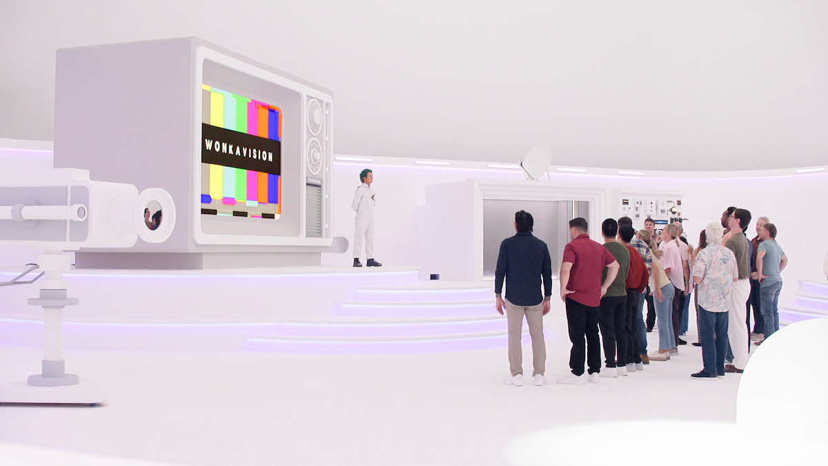 A group of people stands in a futuristic, all-white room watching a person in a white coat near a giant retro TV displaying "WONKAVISION" and color bars; high-tech equipment and modern lighting fill the space.