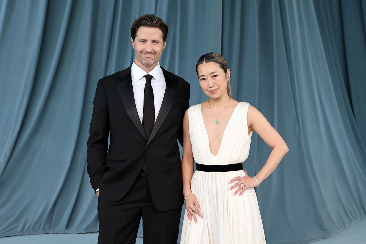 A man in a black tuxedo and a woman in a white gown with a black belt pose together, standing on a light blue carpet in front of draped blue fabric at a formal event.
