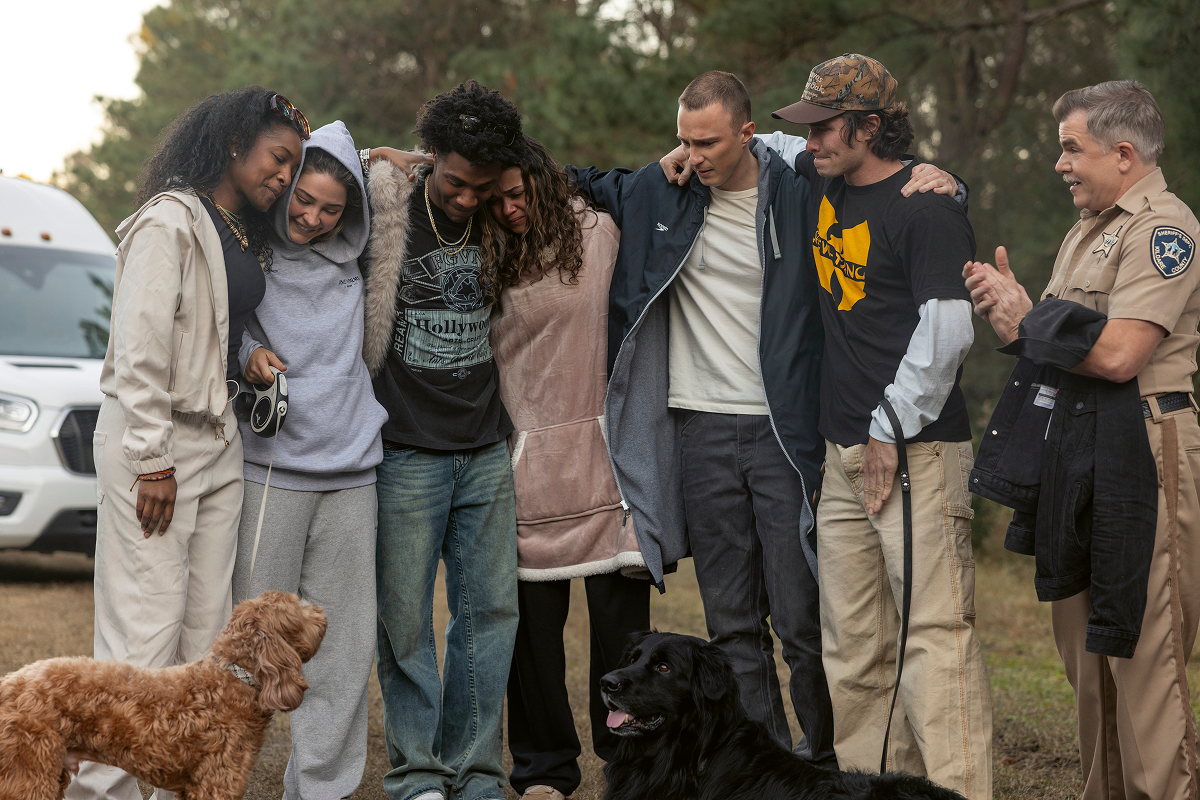 A group of six people and two dogs standing outdoors, some with arms around each other, near a police officer and van, in a wooded area, sharing a friendly, relaxed moment.