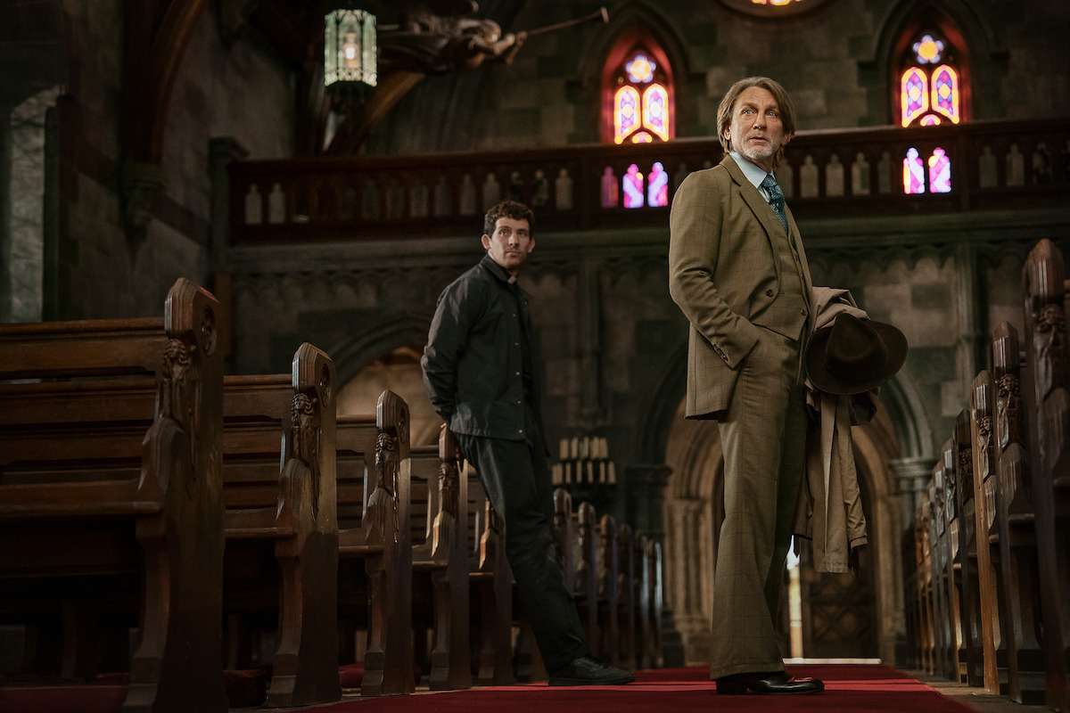 Two men stand in the aisle of an old, dimly lit church with stained glass windows. One is dressed formally in a suit holding a hat, the other is in casual dark clothing. The mood is tense and contemplative.