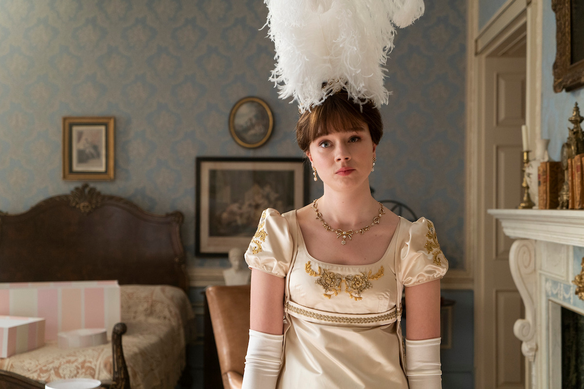 Young woman in an ornate cream gown with gold details and a large feathered headpiece stands in an elegant vintage bedroom with framed art, a fireplace, and a canopy bed.