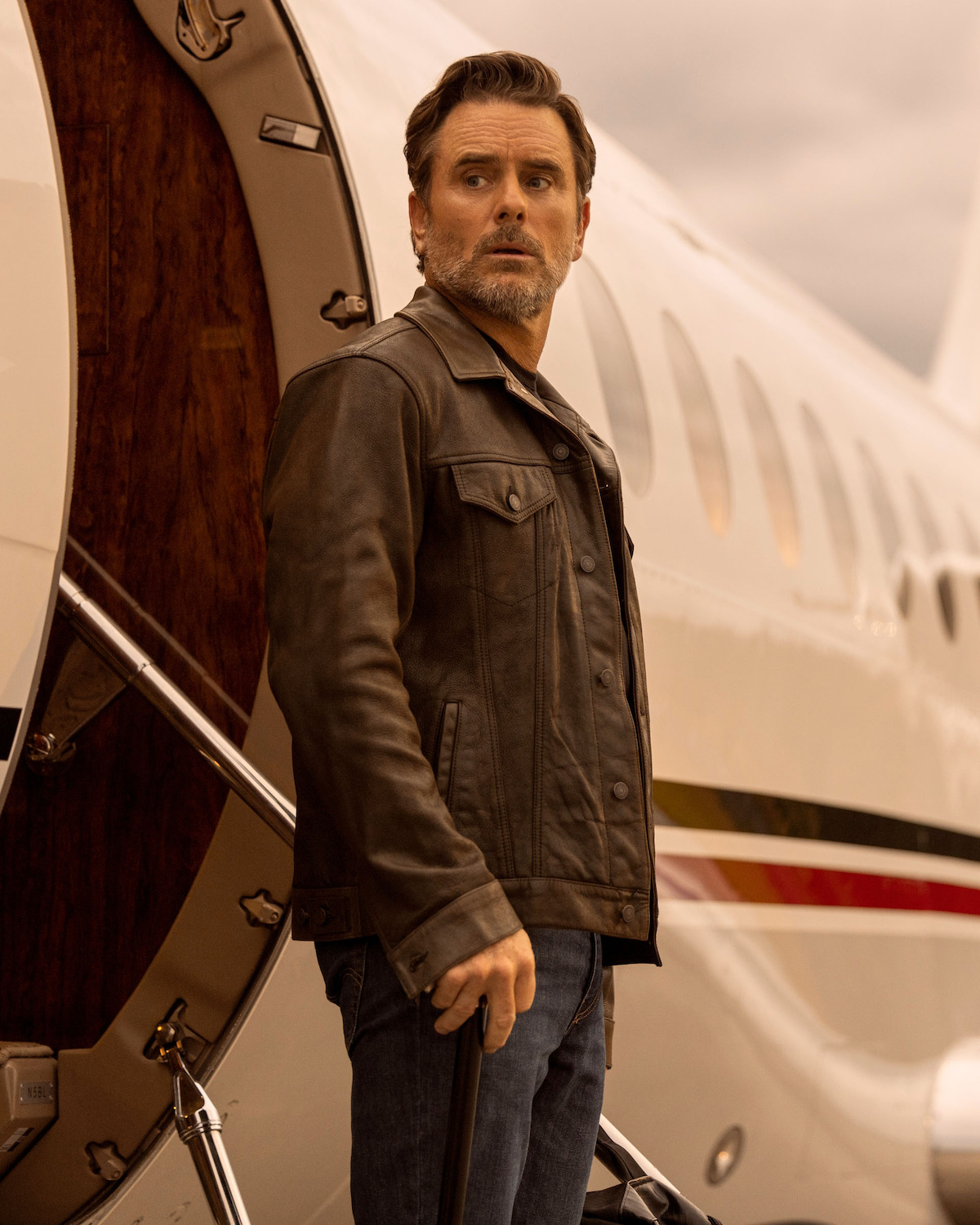 Charles “Chip” Esten as Ward Cameron, in a leather jacket stands by the open door of a private jet, looking to the side with a serious expression under a cloudy sky, creating a tense and dramatic atmosphere.