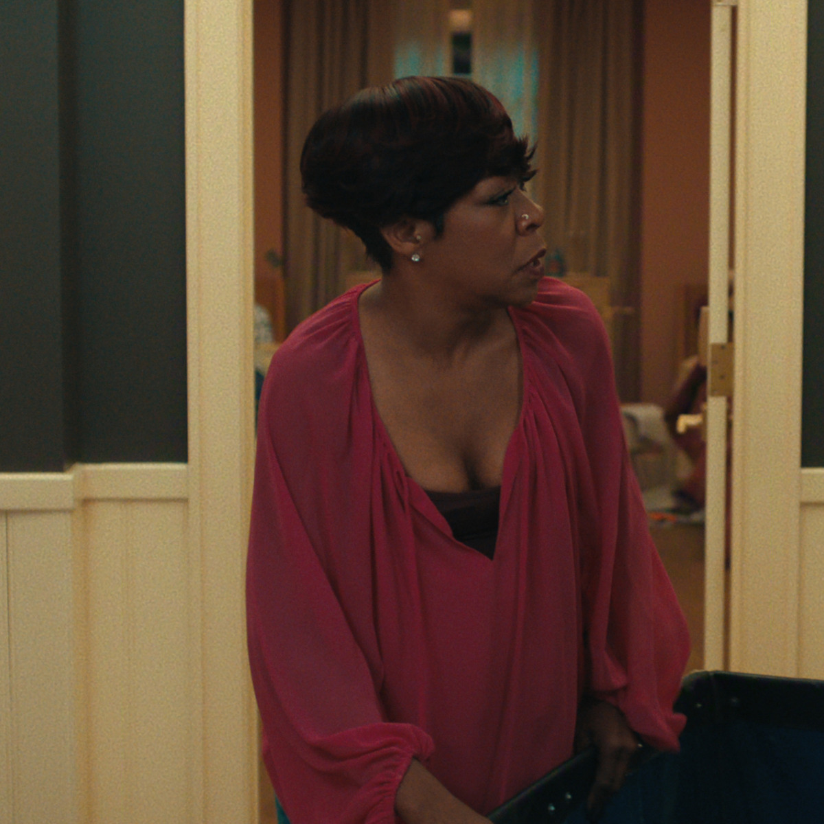 Woman in a deep pink blouse stands indoors, looking to her left, holding an object. The setting appears to be a warmly lit room with cream paneled walls, an open door, and a glimpse of a furnished living area behind her.