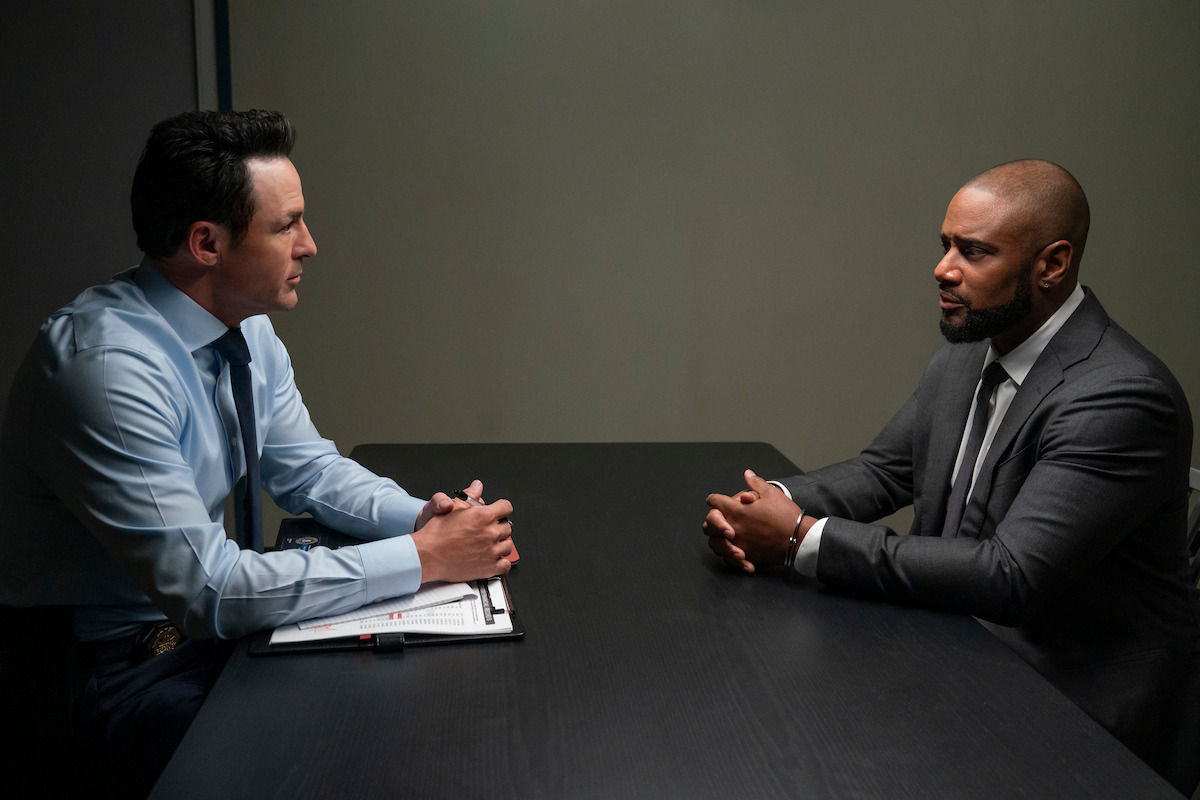 Two men in formal attire sit across a dark table in a serious discussion in a plain, dimly lit room, suggesting an interrogation or interview setting with a notepad and pen on the table.