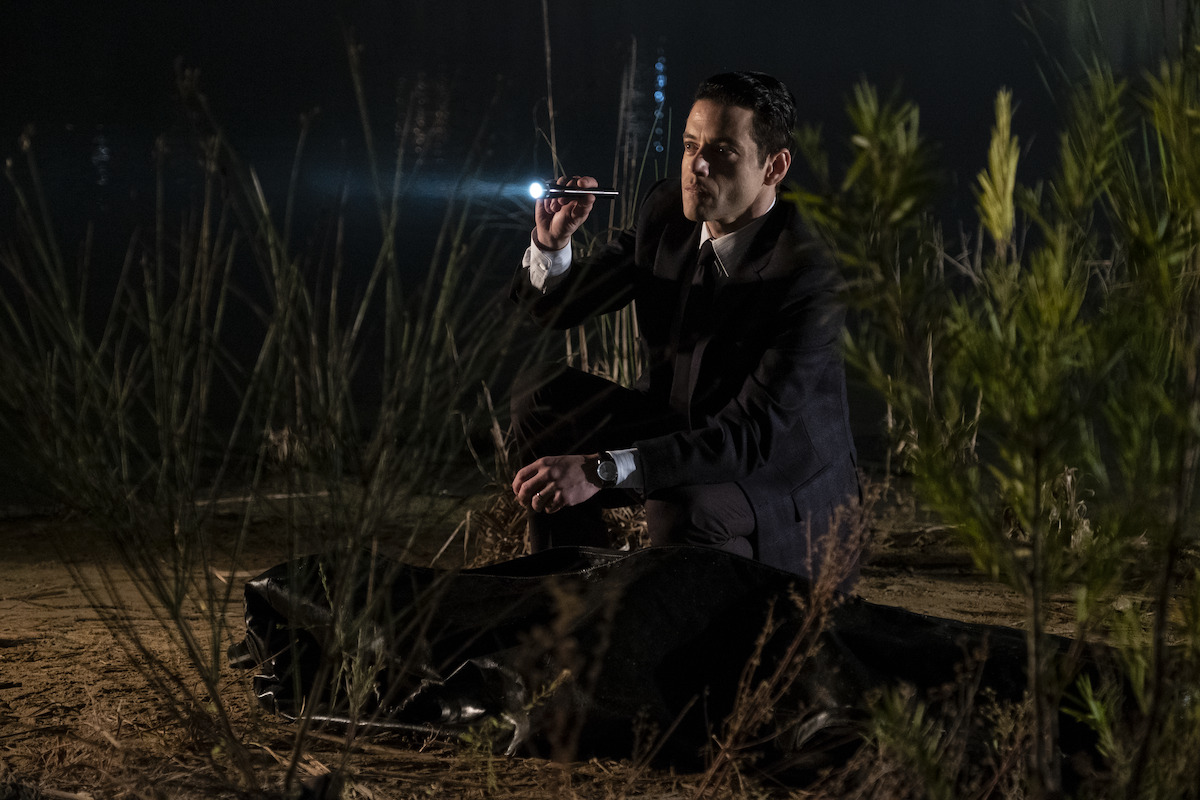Rami Malek crouches down amid foliage in the dark and shines a flashlight in a still image from ‘The Little Things.’