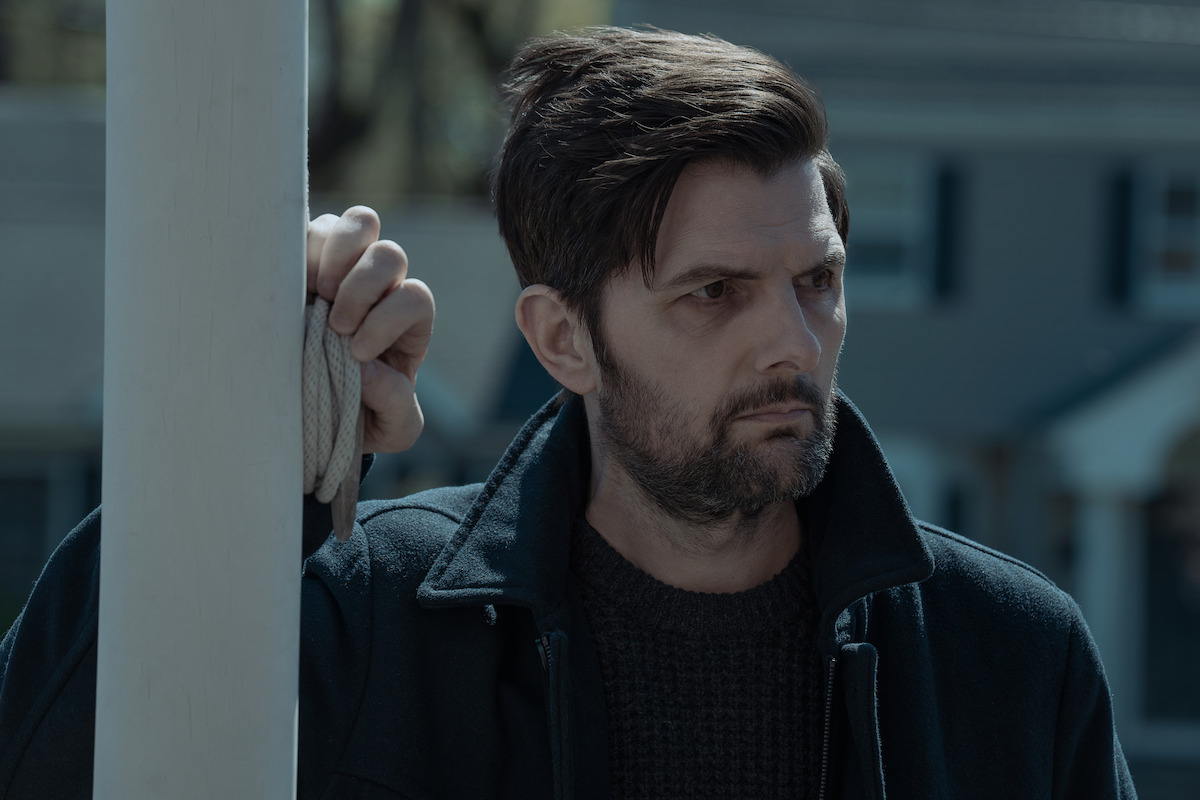 Bearded man in dark clothing stands outdoors, gripping a white pole, with a serious expression. Suburban house with blue siding and windows appears blurred in the background, suggesting a residential neighborhood setting.