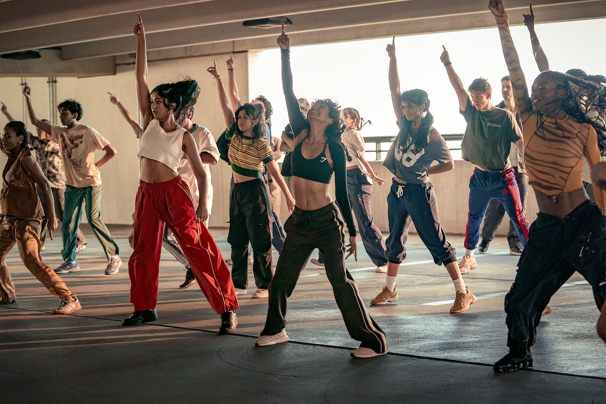 A diverse group of dancers practice a choreographed routine in a sunlit, urban parking garage, all striking dynamic poses with one arm raised, wearing sporty, casual clothes.