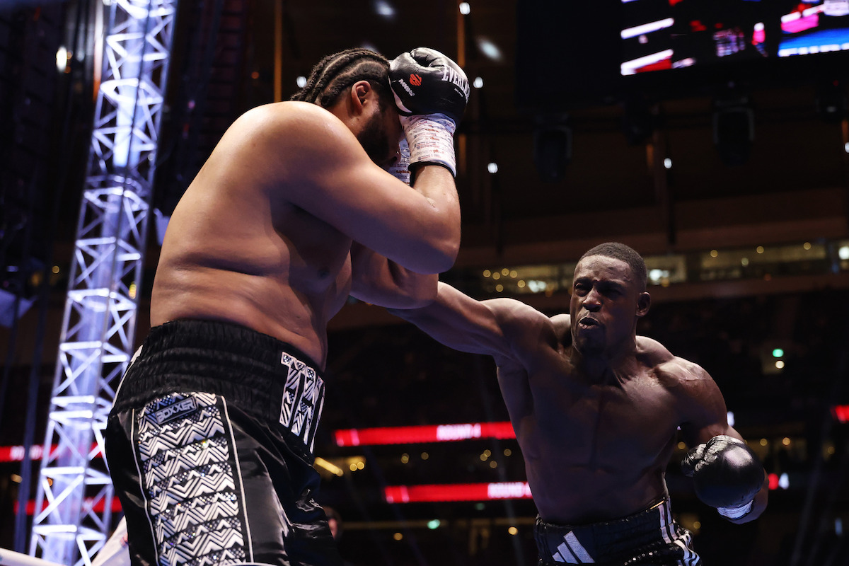 Two boxers in a boxing ring during a match, one throwing a punch at the other under bright arena lights with an audience in the background.