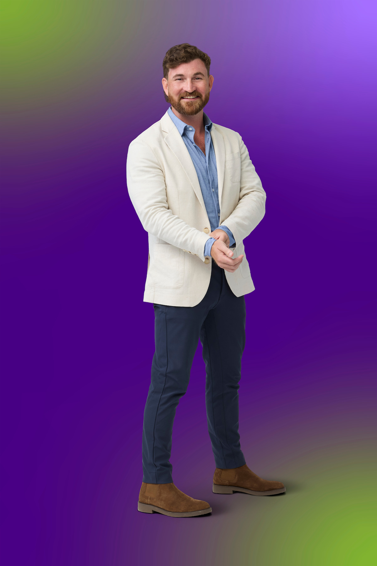 Man with short hair and beard wearing a white blazer, blue shirt, dark pants, and brown shoes, standing and smiling against a vibrant purple and green gradient background.