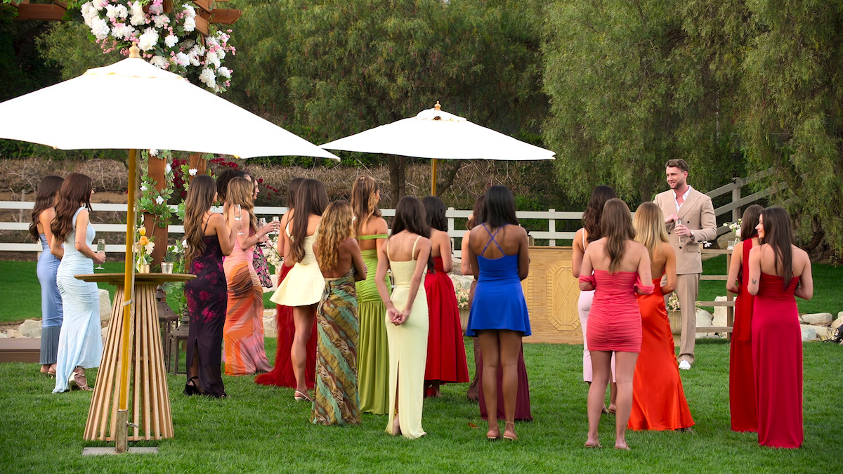 A group of women in colorful dresses stand on grass under white umbrellas, facing a man in a suit outdoors. The scene appears to be a formal gathering or ceremony in a garden or park setting with trees and floral decorations.