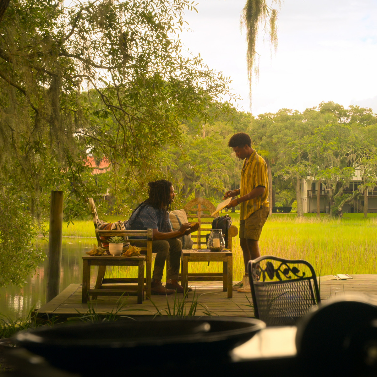 Duas pessoas conversando em um deque de madeira cercado de plantas exuberantes. O deque tem bancos, uma mesa e comida. Um rio ou pântano e várias casas estão visíveis ao fundo sob um céu nublado, dando um clima tranquilo ao ar livre.