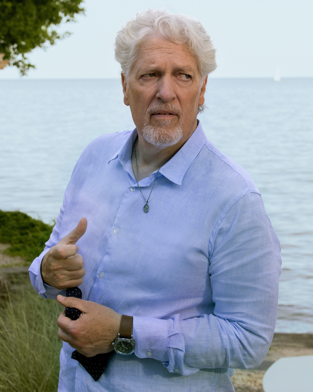 Older man with white hair and beard stands outdoors by a lakeshore, wearing a light blue shirt and holding a necktie. Natural background with rocks, trees, and water on a clear day.