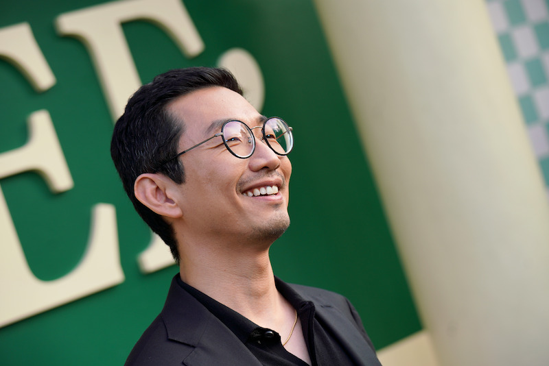Smiling man with glasses in a black suit standing outdoors in front of a green and white wall with large letters, appearing at an event or gathering.