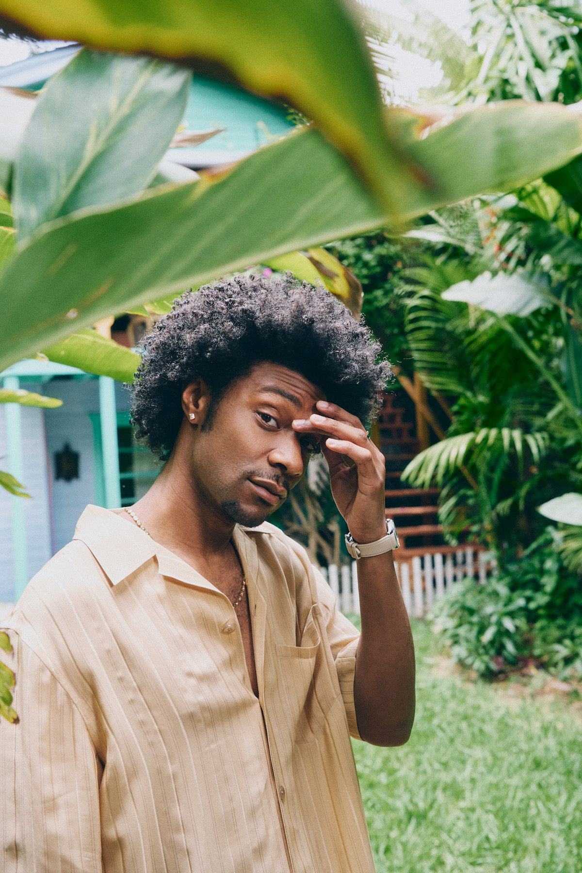 Man with Afro hairstyle standing outdoors in a lush green garden, wearing a beige shirt, touching his forehead, with tropical plants and a house with a picket fence in the background.