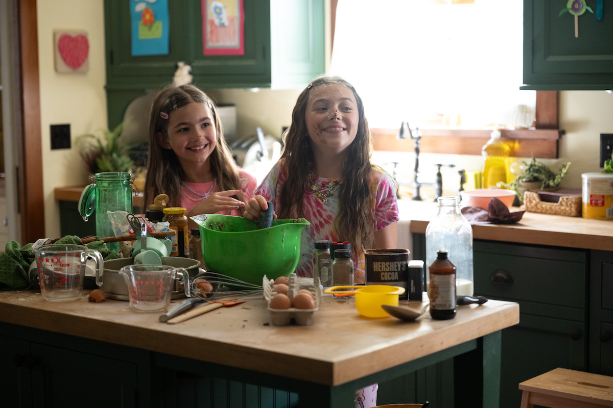 Two smiling girls baking together in a cozy, sunlit kitchen with green cabinets, surrounded by baking ingredients and utensils, including eggs, measuring cups, cocoa, and mixing bowls on the countertop.