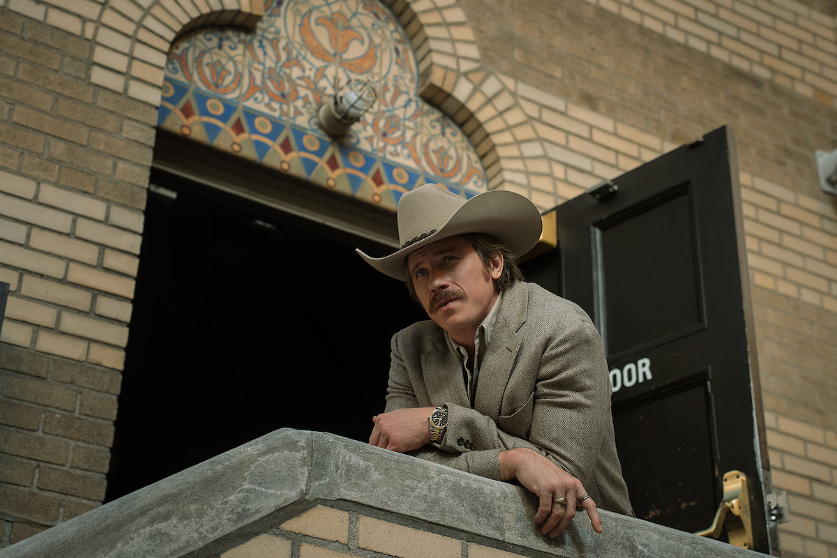 Man in a cowboy hat and suit leans on a concrete railing outside a brick building with decorative arch, open door and ornate tilework above. Daylight scene, western style clothing, casual pose.