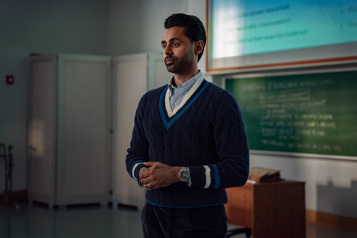 Man standing in a classroom wearing a dark sweater, with a chalkboard and projection screen behind him, giving the appearance of a teacher or lecturer in a school or university setting.