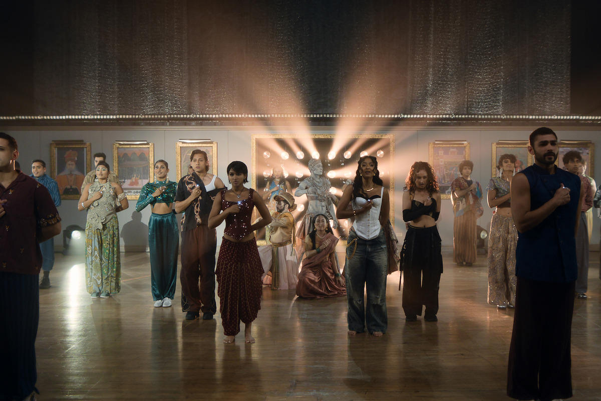 A diverse group of people in artistic clothing stand and pose in a spacious, elegant hall with framed pictures on the walls and dramatic lighting shining from behind a statue, creating a ceremonial, theatrical atmosphere.