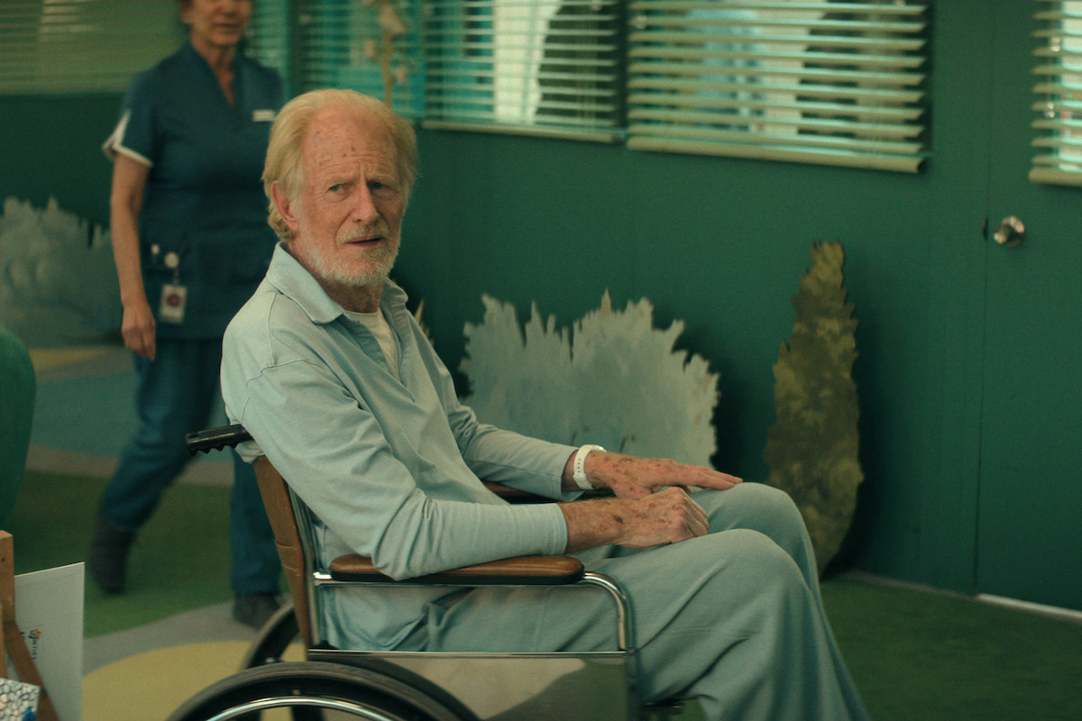 Ed Begley Jr. as Edward Burgen in a wheelchair sits in a medical facility with a nurse standing behind him, surrounded by greenish walls, blinds, and hospital decor.