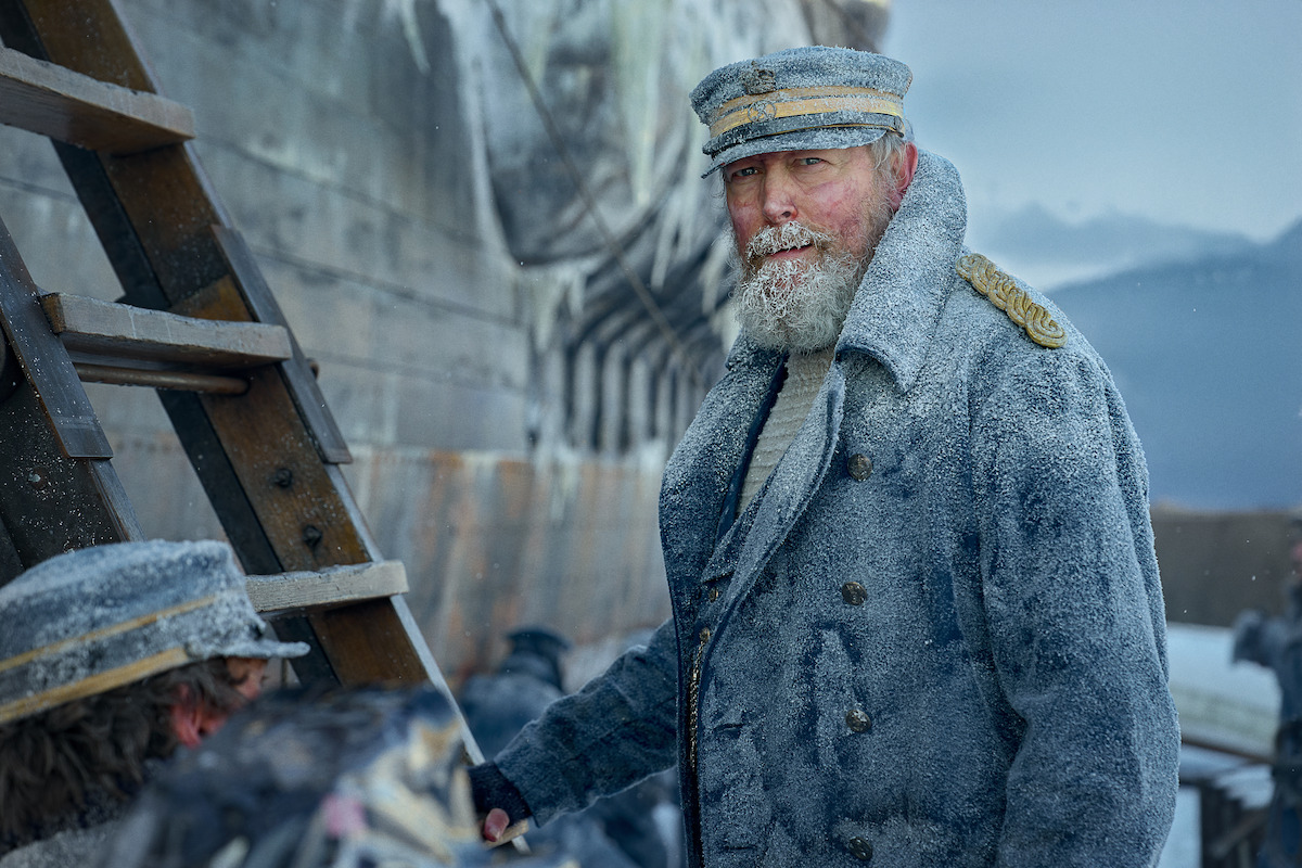 An older bearded man in a frost-covered naval uniform stands outdoors near a ship and wooden ladder, surrounded by icy conditions and crew, creating a bleak, cold, and tense atmosphere.