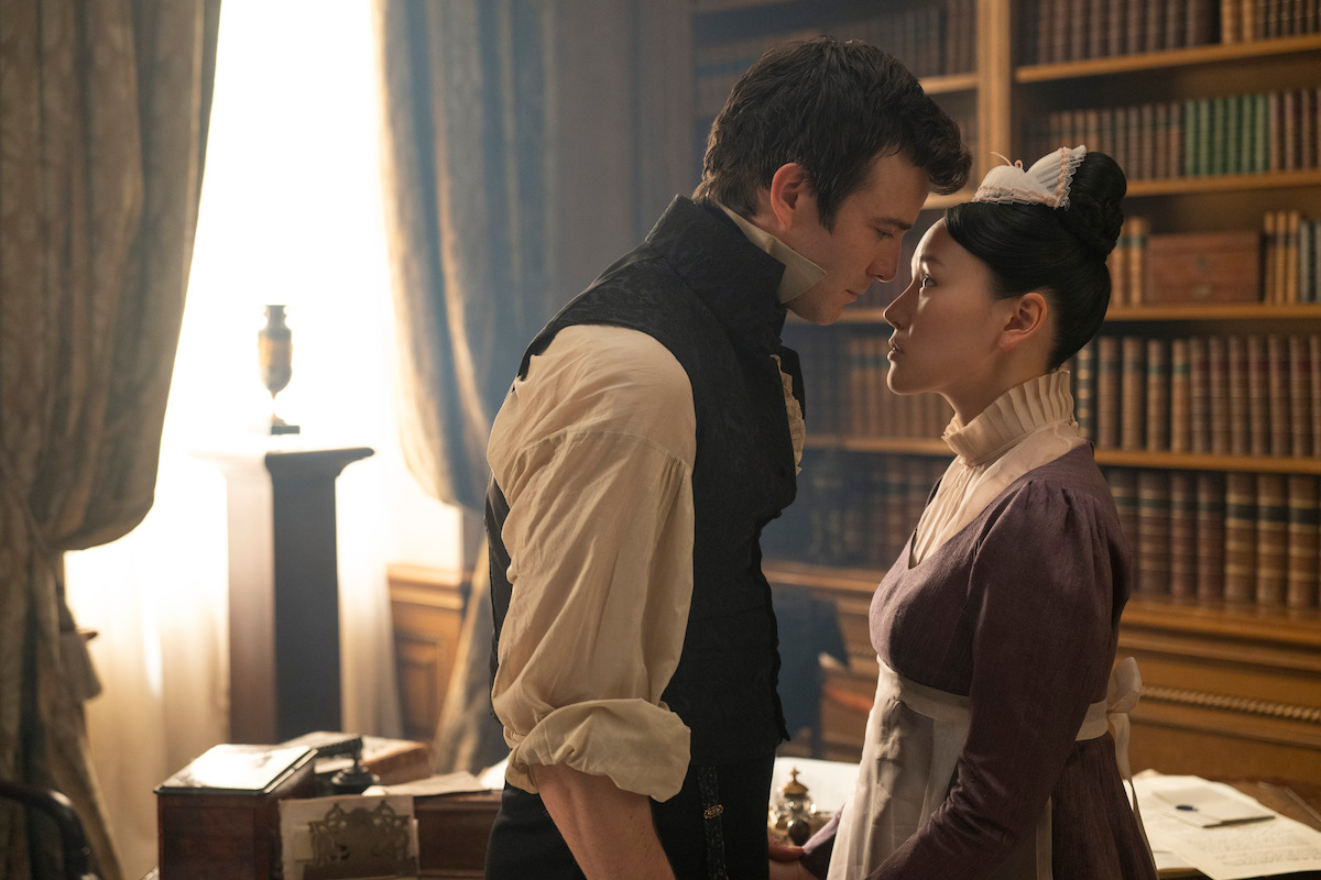 A man and a woman in period costumes stand close together in a library, with shelves of books and a sunlit window in the background, suggesting a tense or intimate moment in a historical setting.