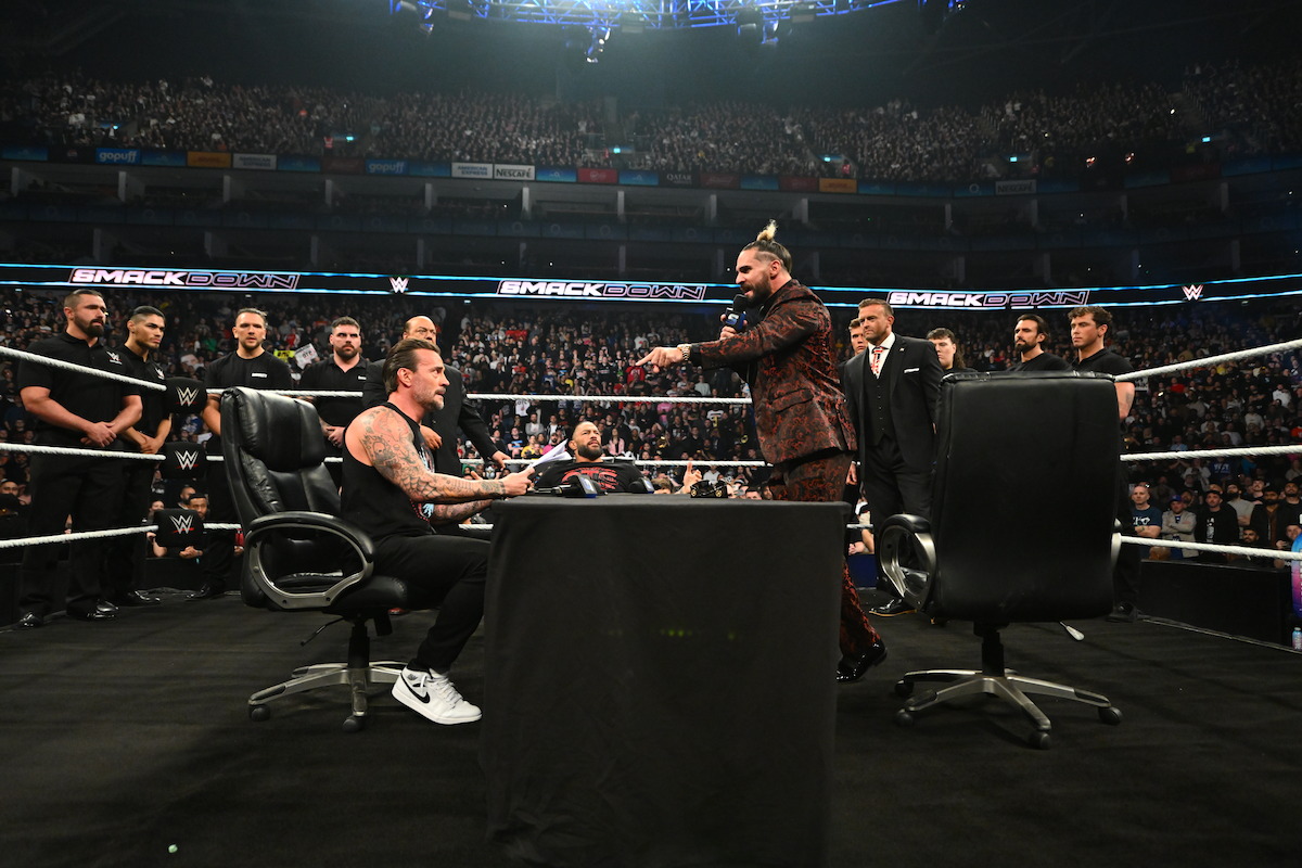 CM Punk, Roman Reigns, and Seth Rollins sit at a desk placed in the ring.