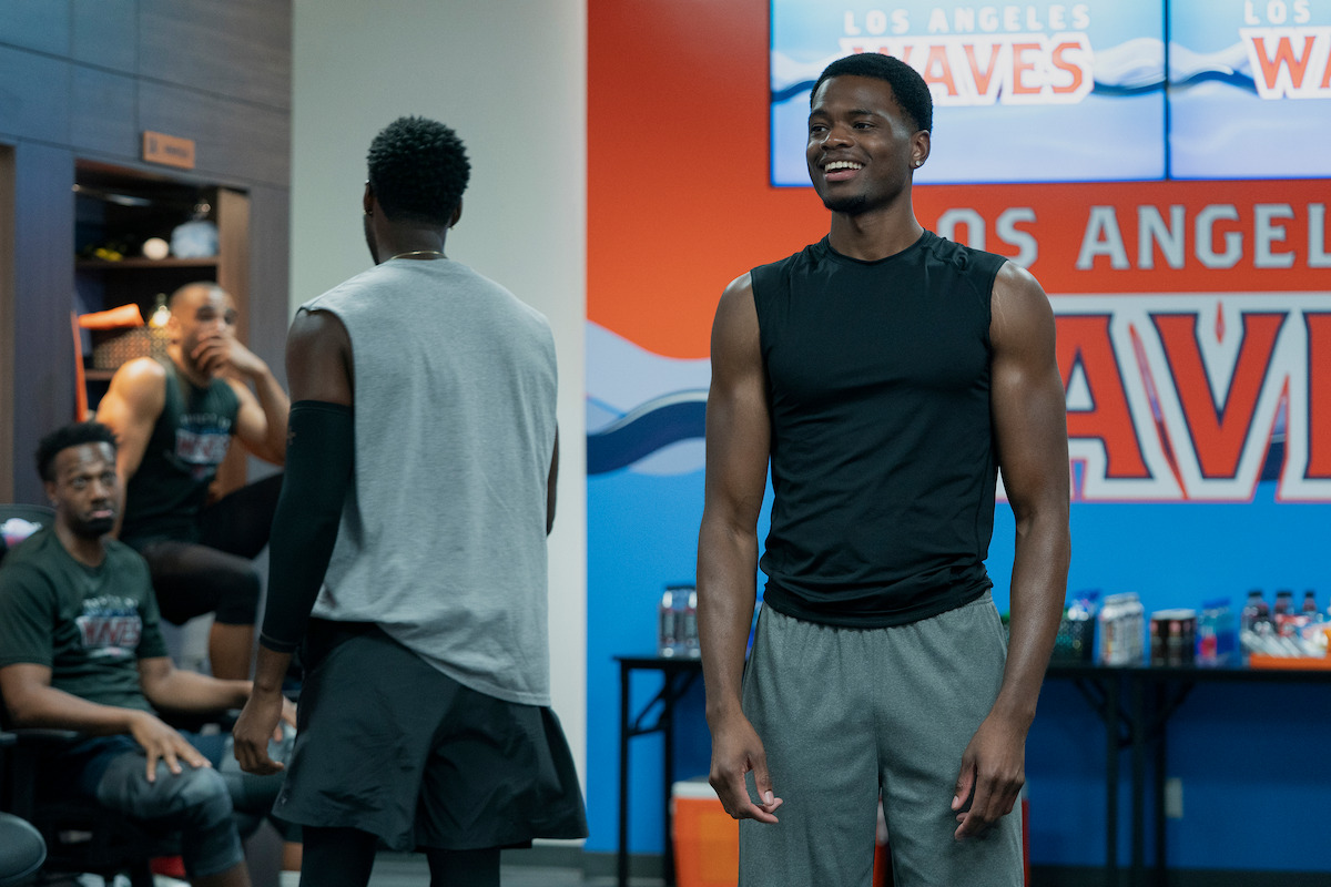 Four men in athletic wear are in a sports team's locker room, with one smiling in the foreground and "Los Angeles Waves" signage and snacks visible in the background.
