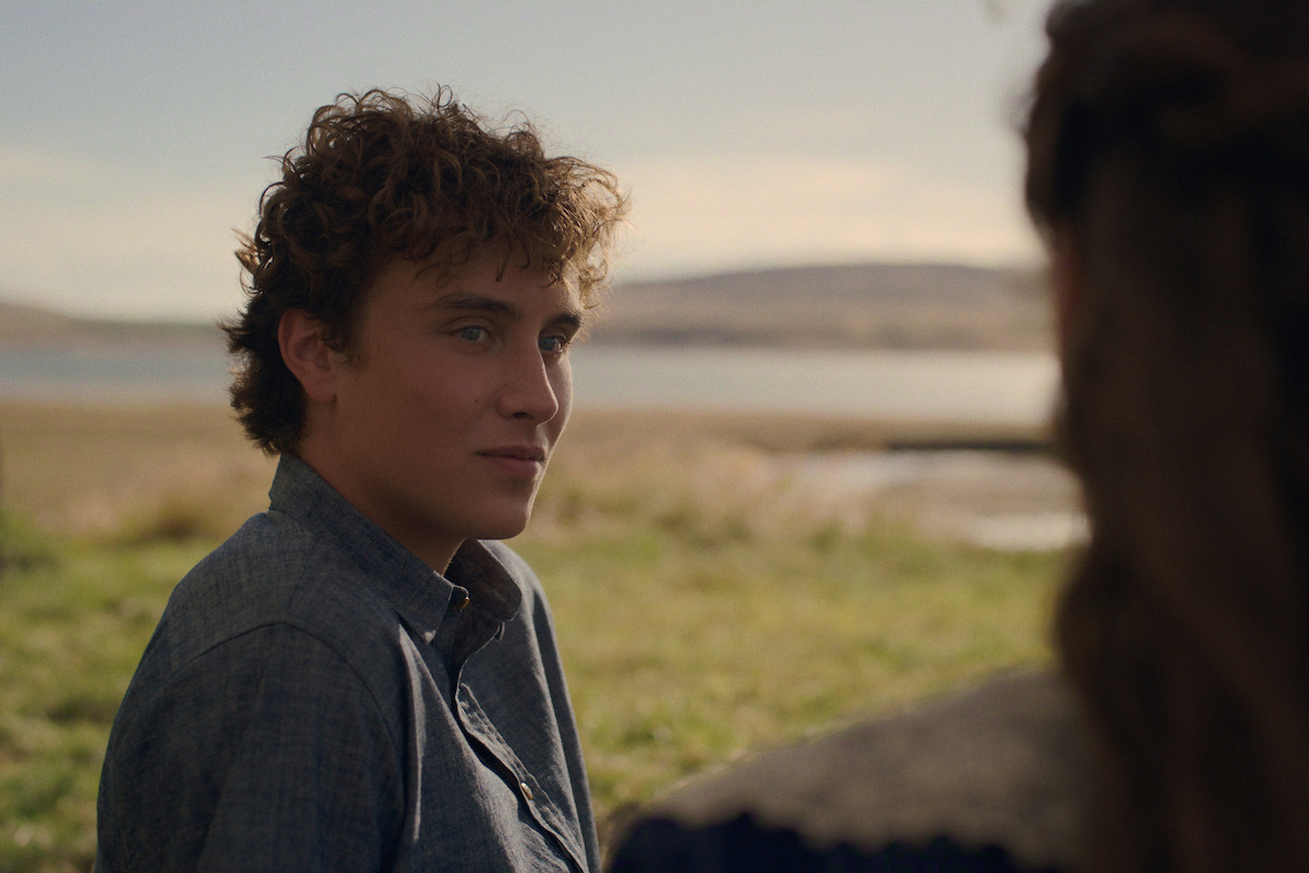 Young man with curly hair outdoors in a grassy field near water, wearing a blue shirt, looking at a person with long hair whose back is to the camera, with hills and a calm sky in the background.